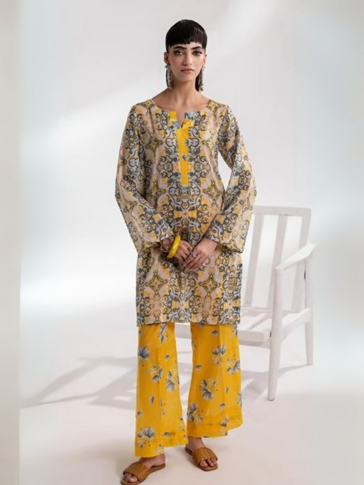 Model wearing a stylish unstitched 2-piece suit featuring a printed shirt with intricate patterns in beige and grey tones, paired with bright yellow floral printed trousers, from Parizad’s premium lawn collect.