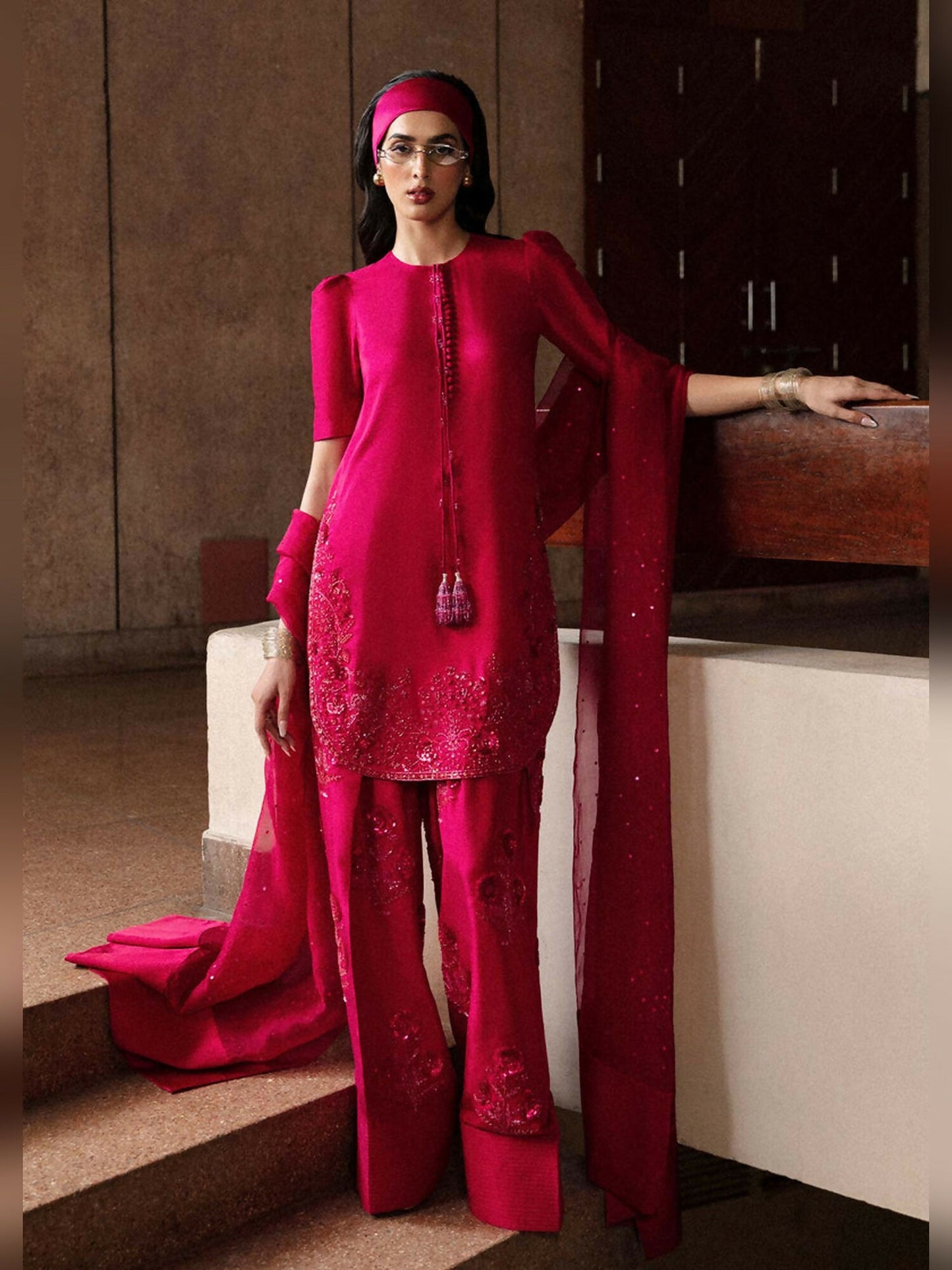Stitched 3-piece suit by Hussain Rehar featuring a hot pink short raw silk shirt with puff sleeves, embellished detailing, and precious stones, paired with an embellished raw silk shalwar, and an organza dupatta with heavy sequin spray and satin silk pallu.