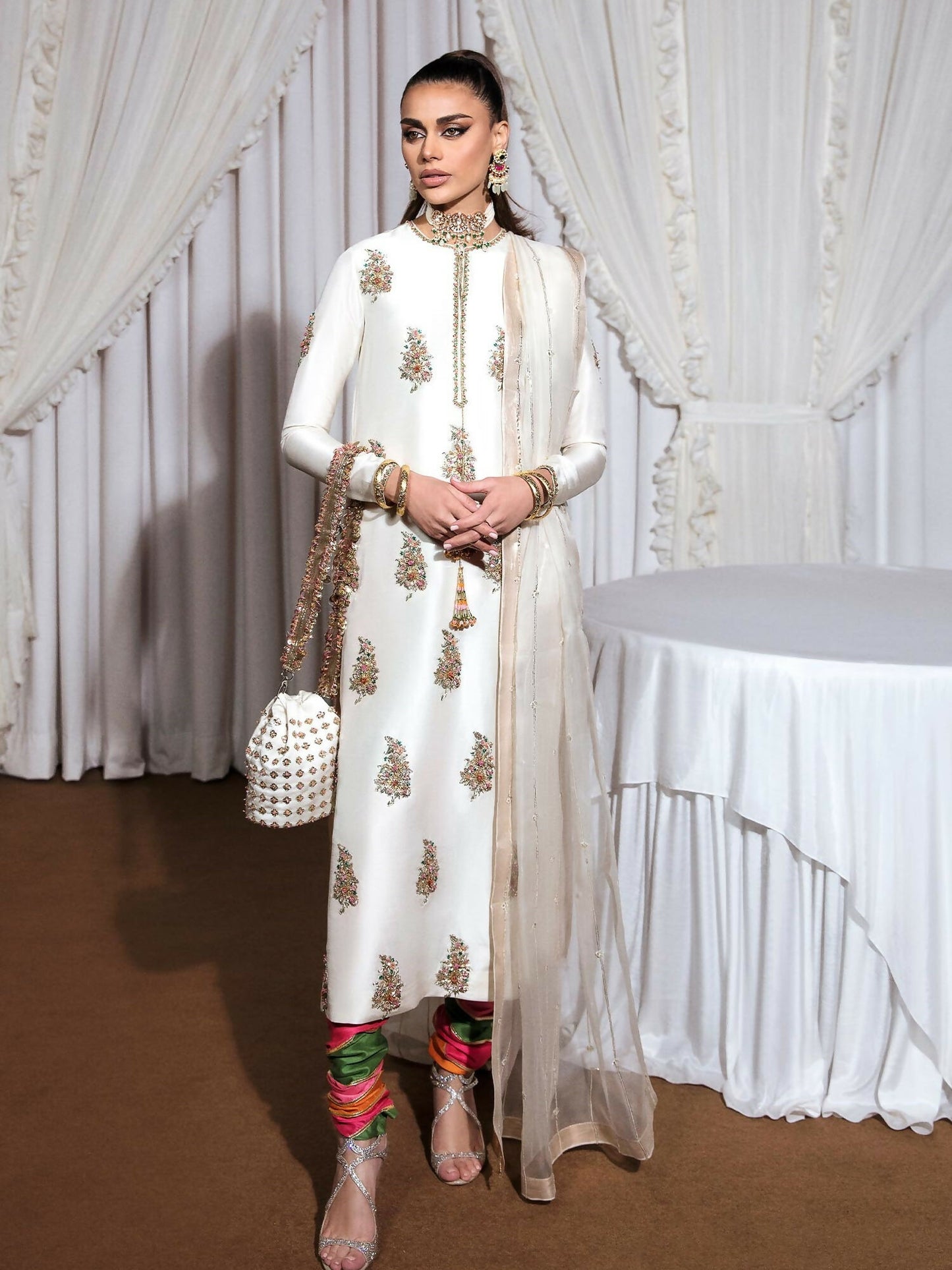 Ivory raw silk stitched 3-piece suit Blanc by HussainRehar featuring an embellished neckline with gold nakshi, sequins, pink crystals, and multicolor bugle beads with tassel details, paired with a multi-colored printed satin chooridar with gotta work and an organza dupatta with multi-head detailing and lappa on all four borders, ideal for festive occasions.