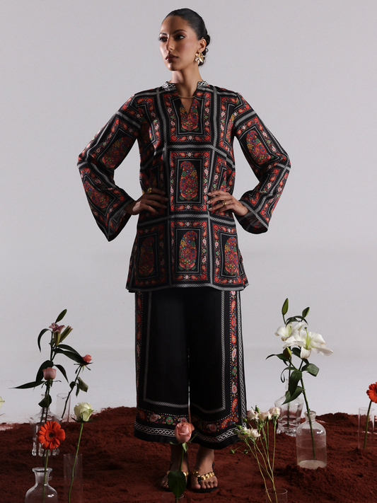 Model in a black digital printed silk co-ord set with colorful framed ethnic motifs on the shirt and bordered wide-leg trousers, standing among scattered flowers against a minimalist backdrop.
