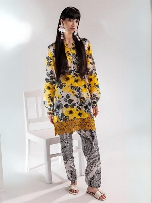 Model wearing an unstitched Parizad 2-piece lawn suit (PA072502) featuring a vibrant yellow floral printed shirt with black and grey accents, finished with a mustard lace hem, paired with white paisley-printed trousers.