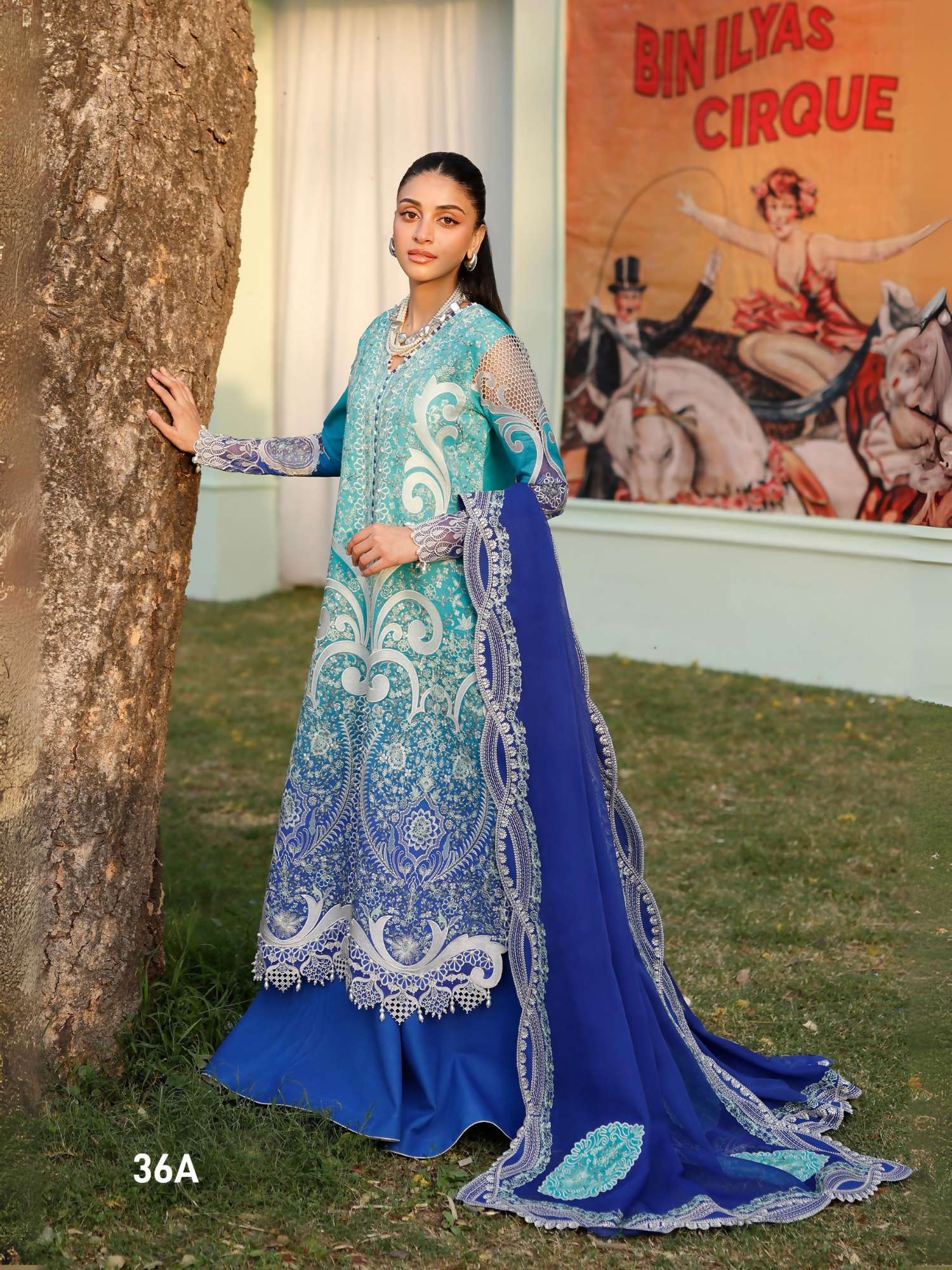 Girl standing wearing unstitched tie & dye printed suit with embroidered front, sleeve motifs, daman lace, and organza detailing, paired with chiffon dupatta featuring embroidered motifs, pallos, and side lace, and dyed cotton trouser.