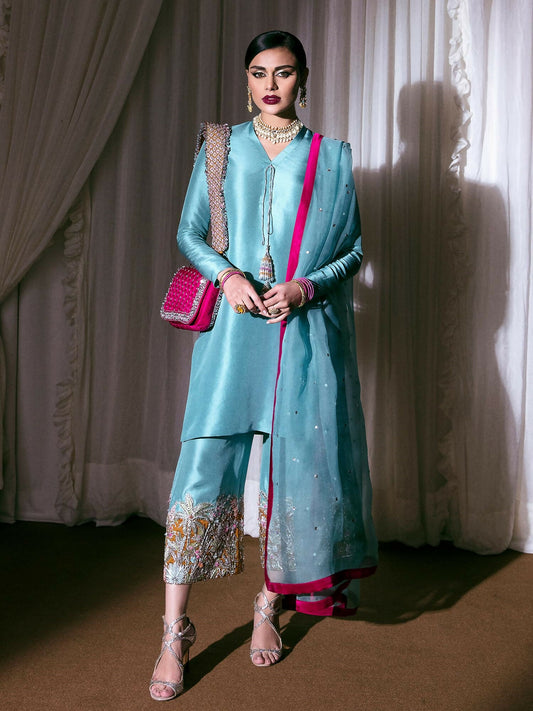 Ice blue raw silk stitched 3-piece suit Azure BNB by HussainRehar featuring a v-neckline shirt with hanging tassel, heavily embellished raw silk culottes with silver nakshi, sequins, dabka, gotta, dibbi, ghorari, multicolor bugle beads and orange piping, paired with a matching organza dupatta adorned with silver sequins and satin silk borders, perfect for festive occasions.