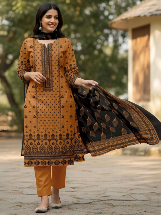 A woman in a mustard printed suit with a black leaf-patterned dupatta and matching trousers, smiling while standing on a stone path with greenery in the background.