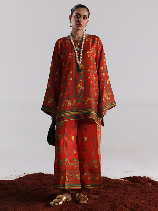 Model wearing a vibrant red digital printed silk co-ord set featuring floral and traditional motifs, with wide-legged trousers and an intricately bordered shirt; accessorized with layered pearls and bold jewelry.